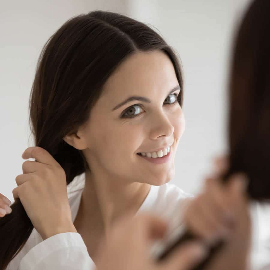A woman with long dark hair smiles while looking in a mirror, holding and styling her hair with both hands. She wears a white top and appears happy and relaxed.