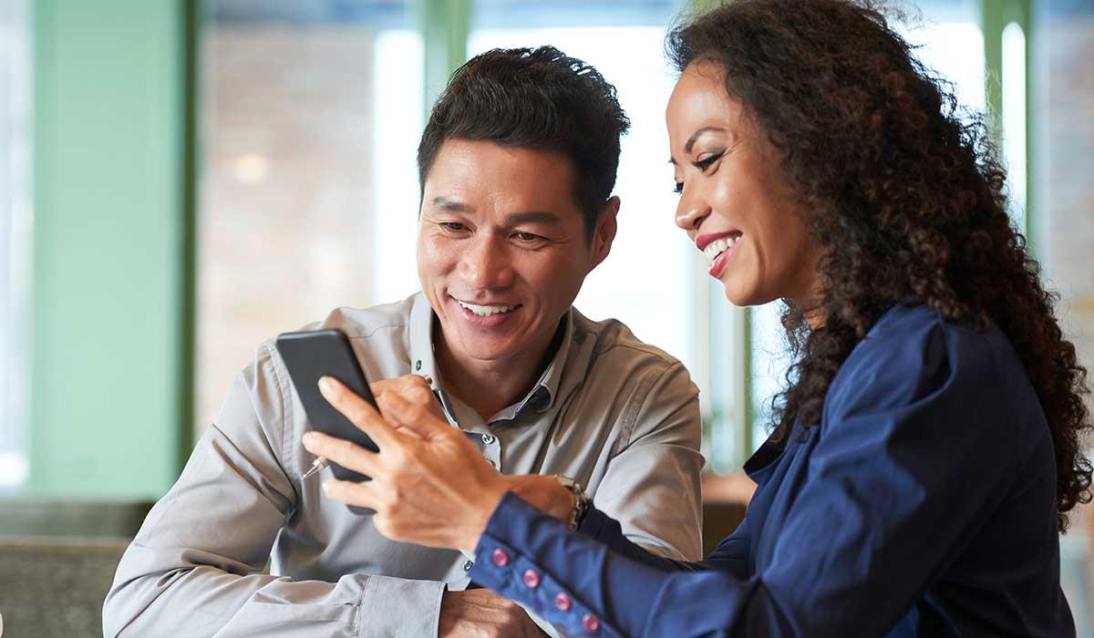 Two people sitting together at a table, smiling and looking at a smartphone that one of them is holding, in a brightly lit indoor setting.