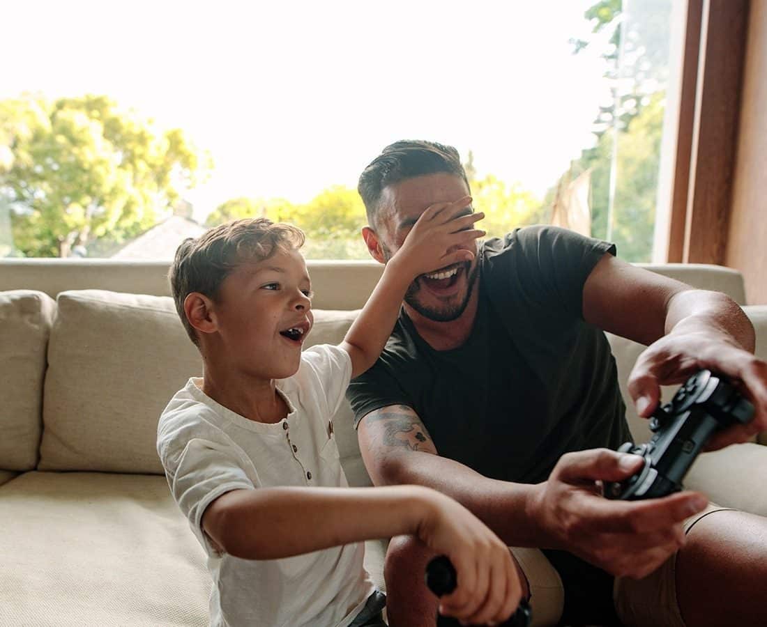 A young boy and a man sit on a couch playing video games. The boy laughs and covers the mans eyes with his hand while the man smiles, holding a game controller. Sunlight streams in from a window behind them.
