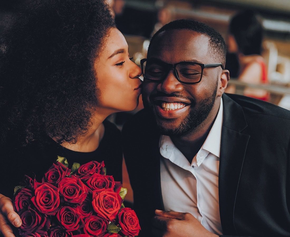 A woman holding a bouquet of red roses kisses a smiling man on the cheek. The man wears glasses and a dark suit, while the woman has curly hair. They appear happy and are sitting close together.