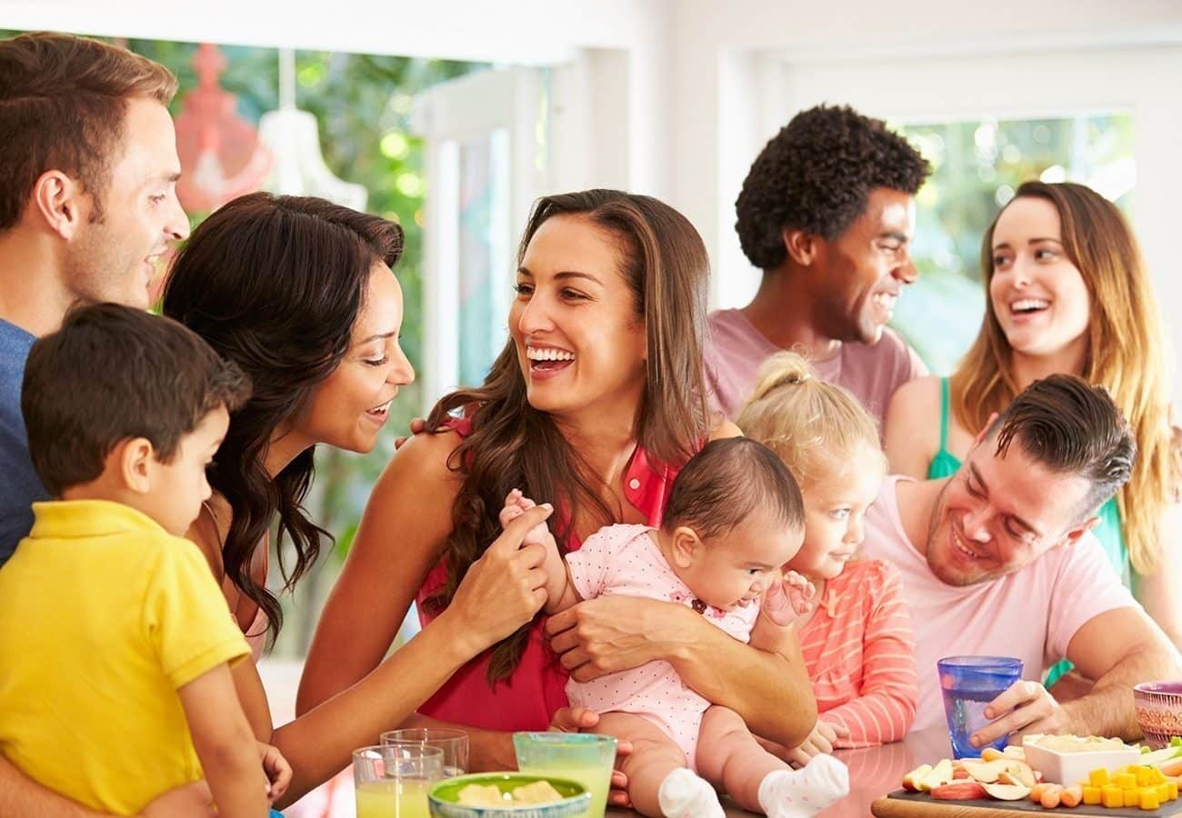 A group of adults and children gather around a table with food and drinks, laughing and smiling in a bright, cheerful room.