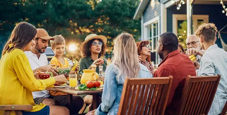 A group of people of various ages sit around a wooden outdoor table, enjoying a meal together. The table has food, drinks, and fresh produce. String lights and greenery create a warm, festive atmosphere.